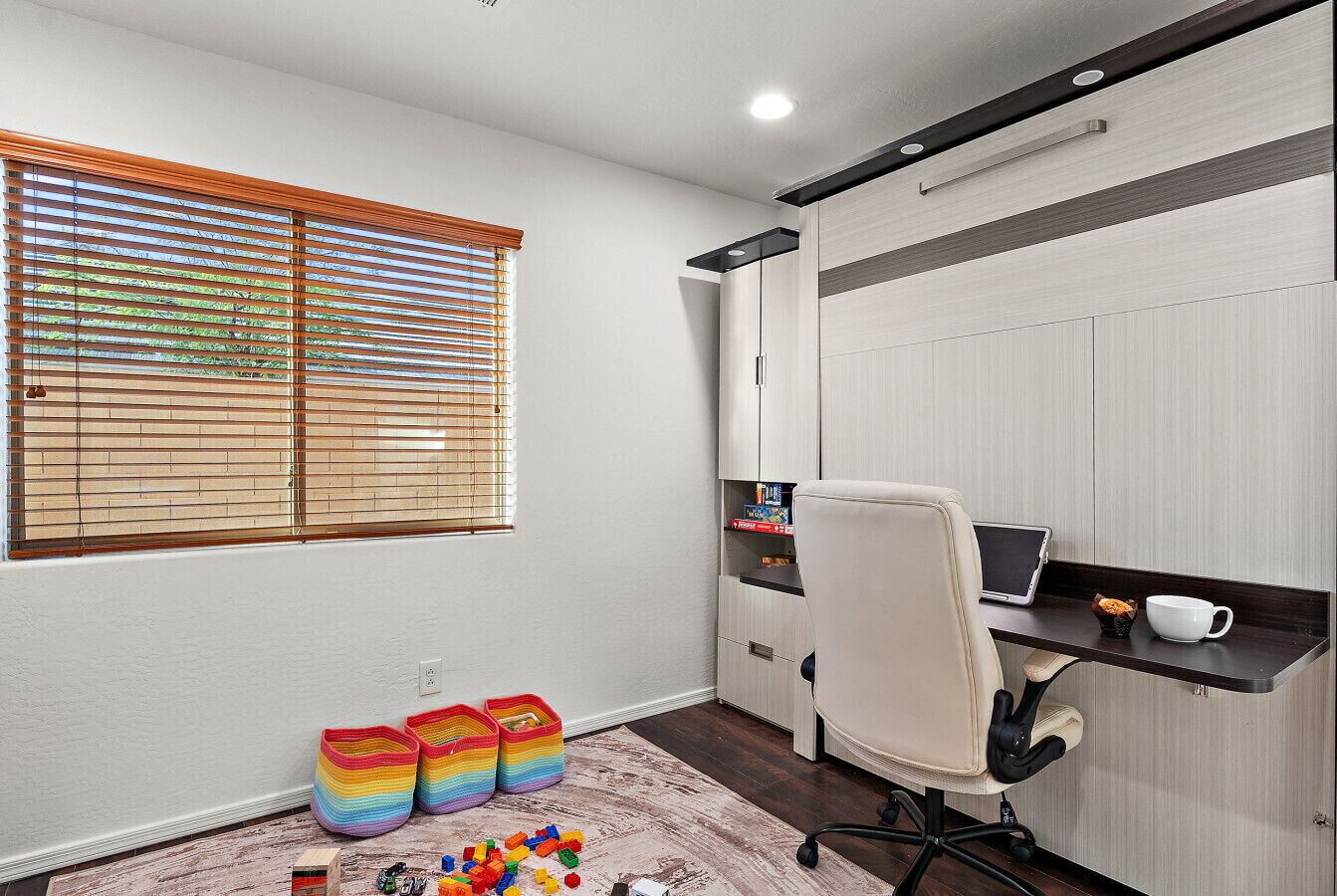 Une pièce comprenant un bureau, une chaise et des armoires de rangement. On y trouve une fenêtre avec des stores, un tapis et des boîtes de rangement colorées.