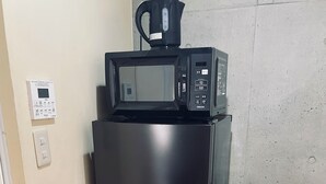 Een zwarte koelkast met een magnetron erboven, een stopcontact en een thermostaat aan de muur.