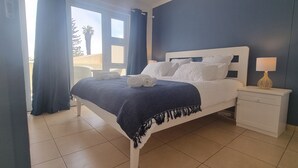 A neatly made bed with a dark blue bedspread and white pillows, a bedside lamp, and a view of the outdoors through a window.