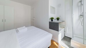 A modern hotel room with a large bed, a built-in wardrobe, a small bathroom with a shower, and a plant on the vanity.