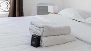 A hotel room with a neatly made bed, a welcome box, a bedside table with a lamp, and a window with curtains.