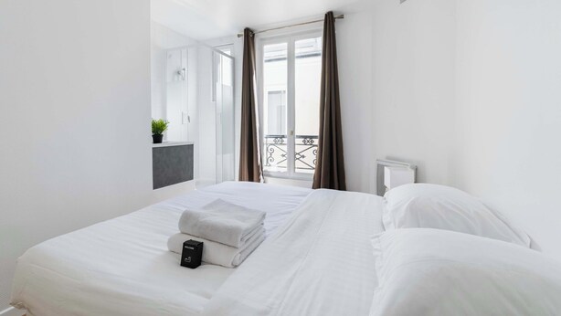 A hotel room with a bed, a bedside table, a window with curtains, and a view of a balcony.
