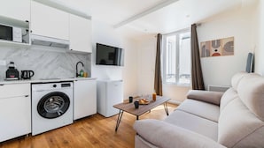 A compact living space with a kitchen, a washing machine, a small table with a bottle and glasses, and a sofa.