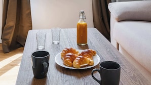 A breakfast table with croissants, juice, and two mugs, a window with curtains, and a wall with abstract art.