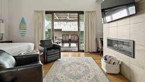 Una habitación de hotel moderna con una cama, una zona de estar con sofá y sillones, una mesa de comedor, un televisor de pantalla plana montado en la pared y un gran ventanal con cortinas.