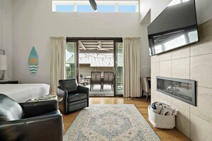 Une chambre d’hôtel moderne comprenant un lit, un coin salon avec un canapé et des fauteuils, une table à manger, un téléviseur à écran plat fixé au mur et une grande fenêtre avec des rideaux.