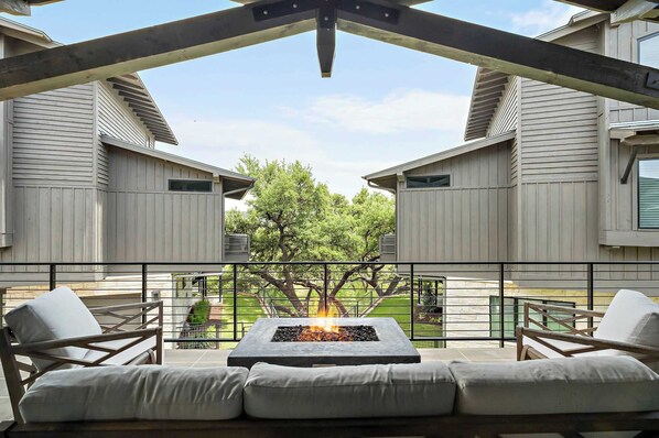 A balcony with a fire pit, outdoor seating, and a view of a tree and building.