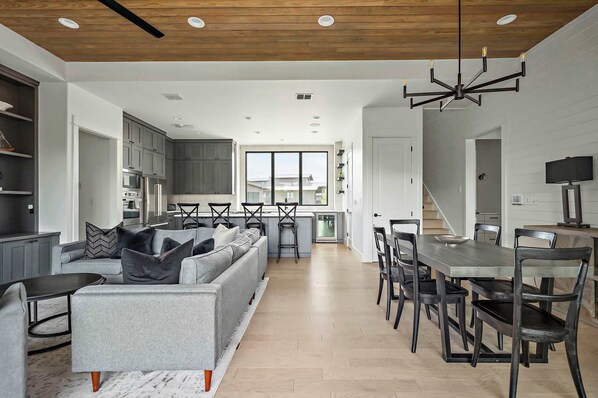 A modern kitchen with grey cabinets, a dining area with a long table and chairs, and a grey sofa with black cushions.