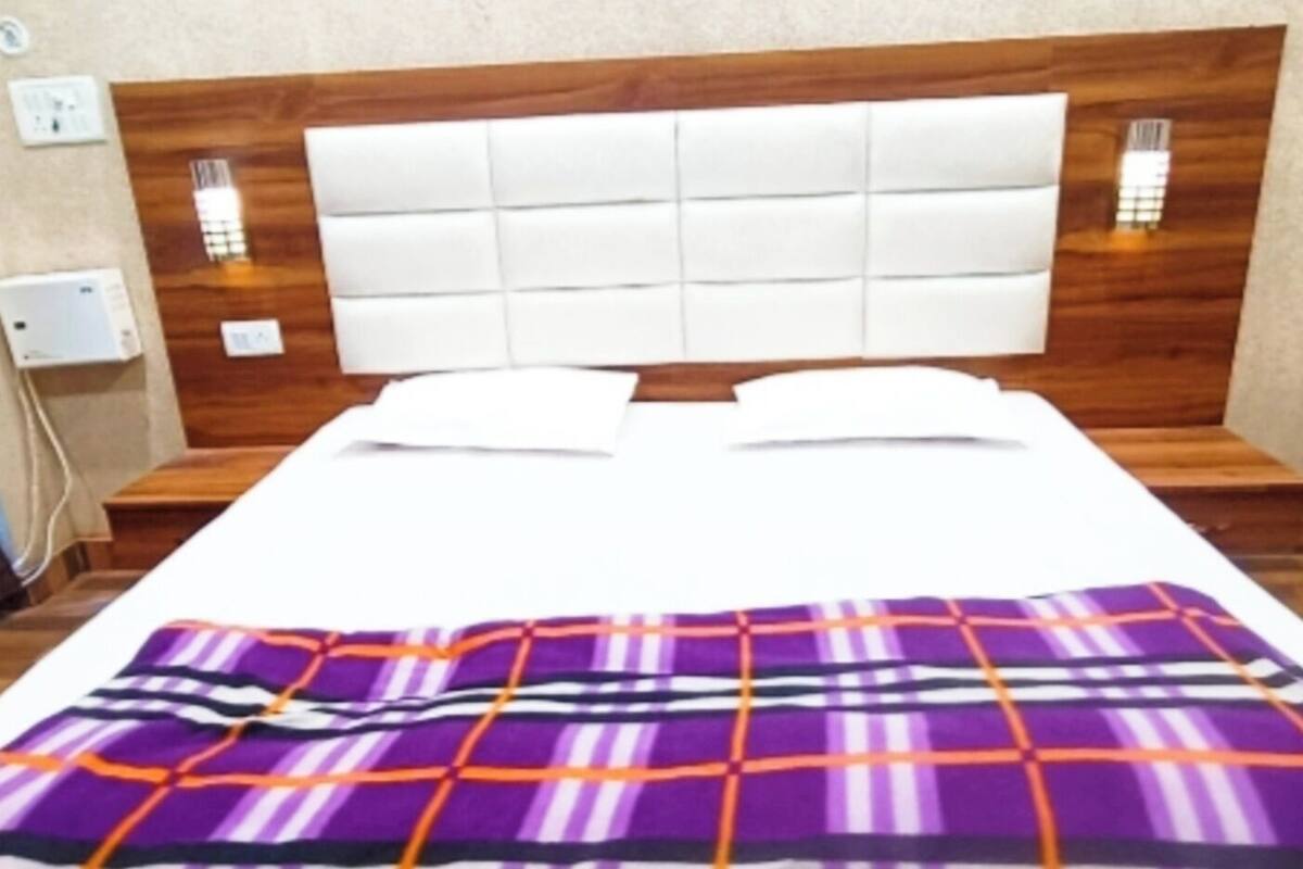 A hotel room with a bed, two pillows, a nightstand, a lamp, and a wall-mounted light.