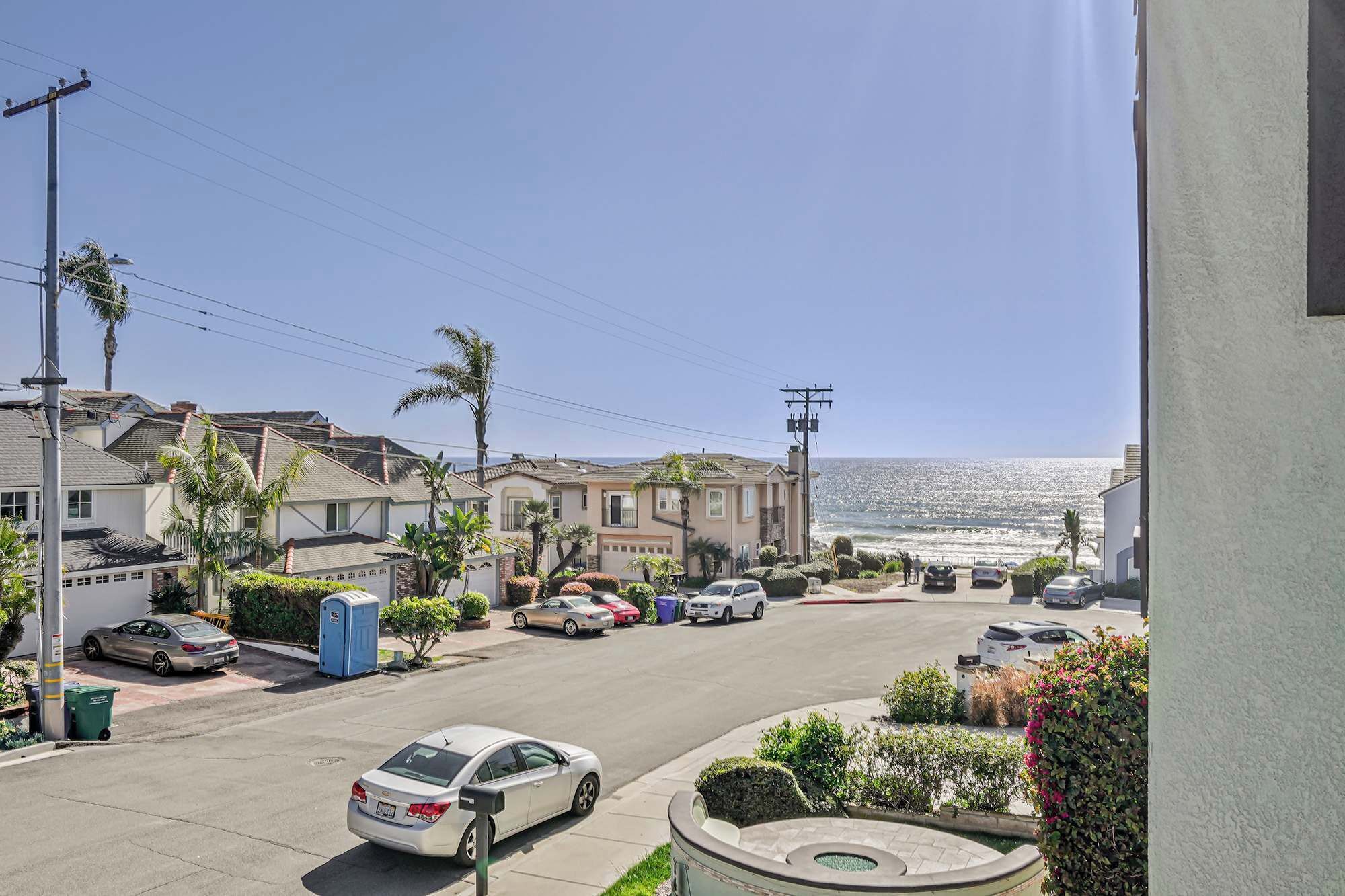 A coastal residential street with houses, parked cars, and a view of the ocean.
