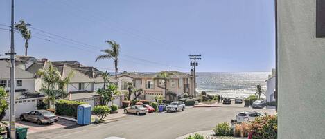 A coastal residential street with houses, parked cars, and a view of the ocean.