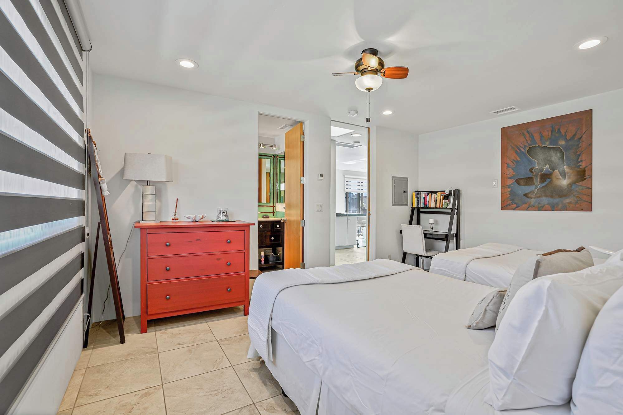 Une chambre avec deux lits, une commode rouge, un ventilateur de plafond et un tableau accroché au mur.
