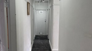 Un pasillo estrecho con paredes blancas y una alfombra gris. Al final del pasillo hay una puerta y una rejilla de ventilación en la pared.