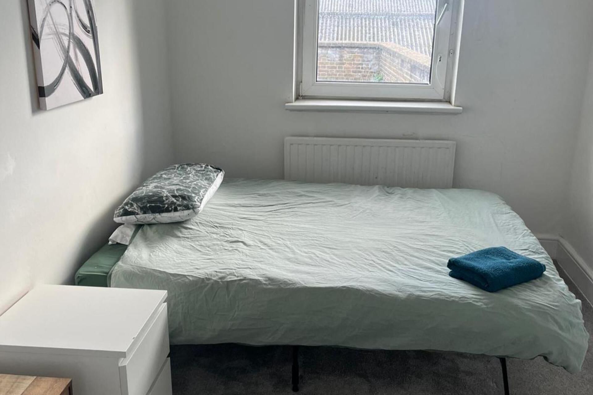 Une petite pièce, peu meublée, avec un lit simple, un radiateur juste au-dessus et une fenêtre donnant sur un mur de briques et une clôture.