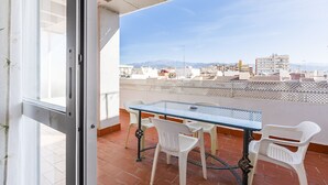 Un balcón con una mesa de vidrio y sillas de plástico blanco, con vistas a un paisaje urbano con edificios y montañas.