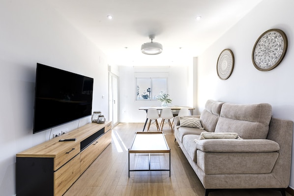 Una sala moderna con televisor de pantalla plana, mueble de entretenimiento de madera, sofá color beige y una zona de comedor con mesa y sillas.