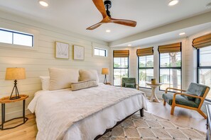 Une chambre à coucher avec un grand lit, un ventilateur de plafond, une chaise, une petite table et une fenêtre avec des stores.