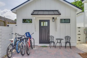 Un bâtiment blanc avec une porte noire, deux fenêtres, un petit patio avec deux chaises et une table, et des vélos garés à l’extérieur.