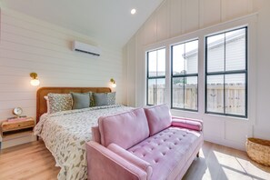 Une chambre à coucher avec un lit, un canapé rose, une table de chevet, un climatiseur fixé au mur et de grandes fenêtres.