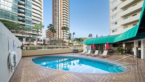 A swimming pool with lounge chairs and umbrellas, surrounded by high-rise buildings and palm trees.