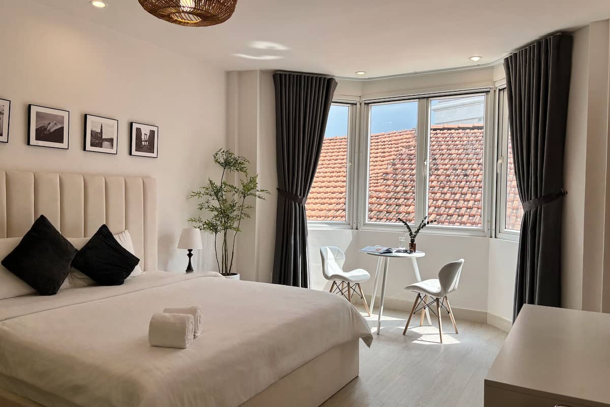 Habitación de hotel con una cama grande, una mesita con sillas, una planta en maceta y vistas a la ciudad a través de la ventana.
