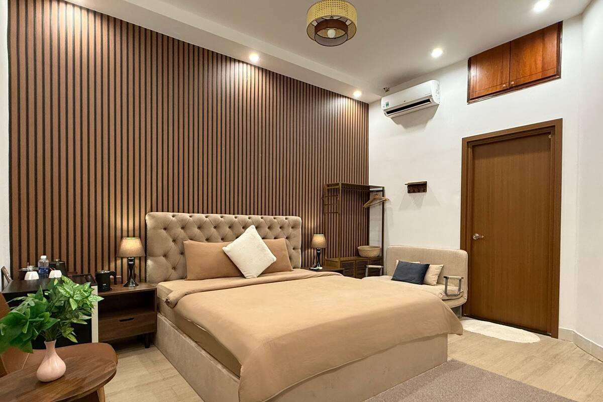 Un dormitorio moderno con un cabecero acolchado, un panel de pared de listones de madera, una mesita de noche con lámpara, una planta pequeña y una puerta de madera.