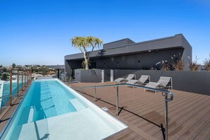 A rooftop pool with a clear view of the city, a modern building with a tree, and lounge chairs.