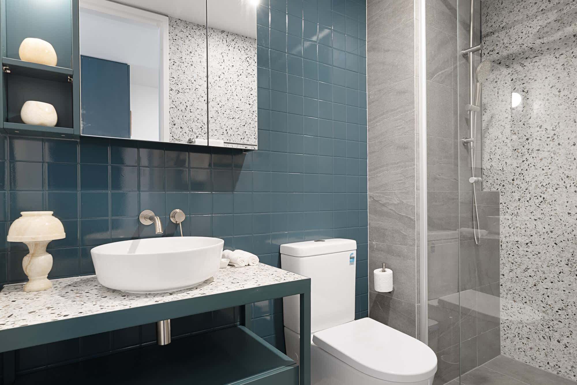 A modern bathroom with a white sink, dark blue tiles, a glass shower enclosure, and a toilet.
