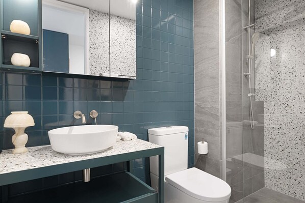 A modern bathroom with a white sink, dark blue tiles, a glass shower enclosure, and a toilet.