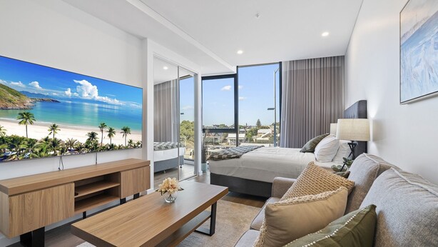 A modern hotel room with a large TV displaying a beach scene, a bed with a view of the beach, a sofa, a coffee table, and a lamp. - Bright Studio in Trendy North Fremantle (North Fremantle)
