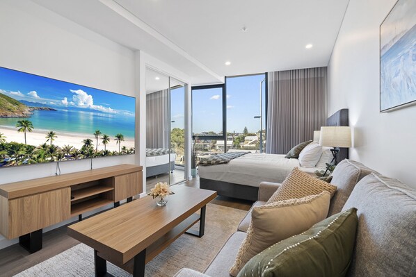 A modern hotel room with a large TV displaying a beach scene, a bed with a view of the beach, a sofa, a coffee table, and a lamp.