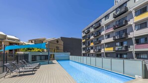 Une piscine entourée de bâtiments à plusieurs étages dotés de balcons, avec des chaises longues et des parasols à proximité.