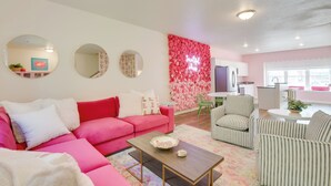 A modern living room with a pink sofa, striped armchairs, and a coffee table. There is a kitchen area in the background with a pink floral wall. - 2 Mi to TBK Bank Sports Complex: Townhome w/ Patio (Bettendorf)