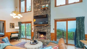 A spacious living room with a stone fireplace, leather sofas, and a large window offering a scenic view. - Sleeps 20! Hot Tub~Outdoor Theater~Fire Pit~Games (Fairview)