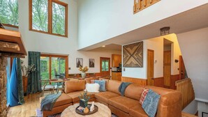 A spacious living room with a large sectional sofa, a round ottoman, and a dining area visible through a glass door. - Sleeps 20! Hot Tub~Outdoor Theater~Fire Pit~Games (Fairview)