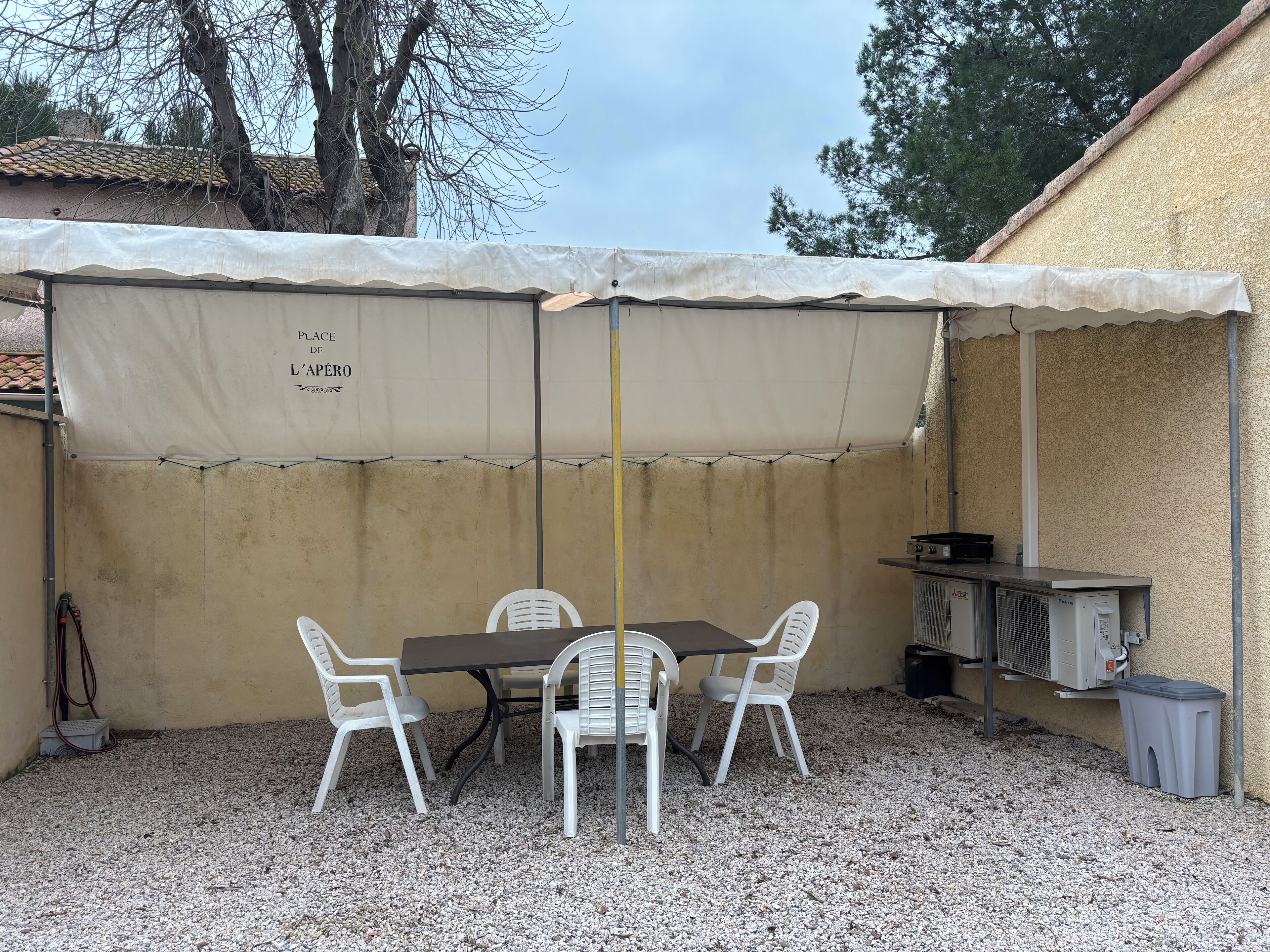 A covered outdoor seating area with a table and chairs, a grill, and a trash bin.