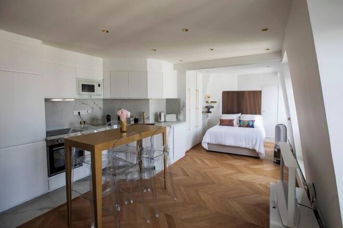 A modern kitchen with a wooden table and clear chairs, a bed with white bedding, and a herringbone floor pattern.