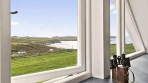 A room with a large window offering a coastal view, a wooden door, and a metal object on the floor. - Swn y Môr - 1 Bedroom Cottage - Whitesands (Saint David's and the Cathedral Close)