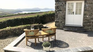 A stone building with a white door, a paved patio with two wooden chairs and a table, and a view of the landscape. - Swn y Môr - 1 Bedroom Cottage - Whitesands (Saint David's and the Cathedral Close)