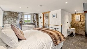 A bedroom with a stone wall, a bed with pillows, a bedside table, and a window with curtains. - Swn y Môr - 1 Bedroom Cottage - Whitesands (Saint David's and the Cathedral Close)