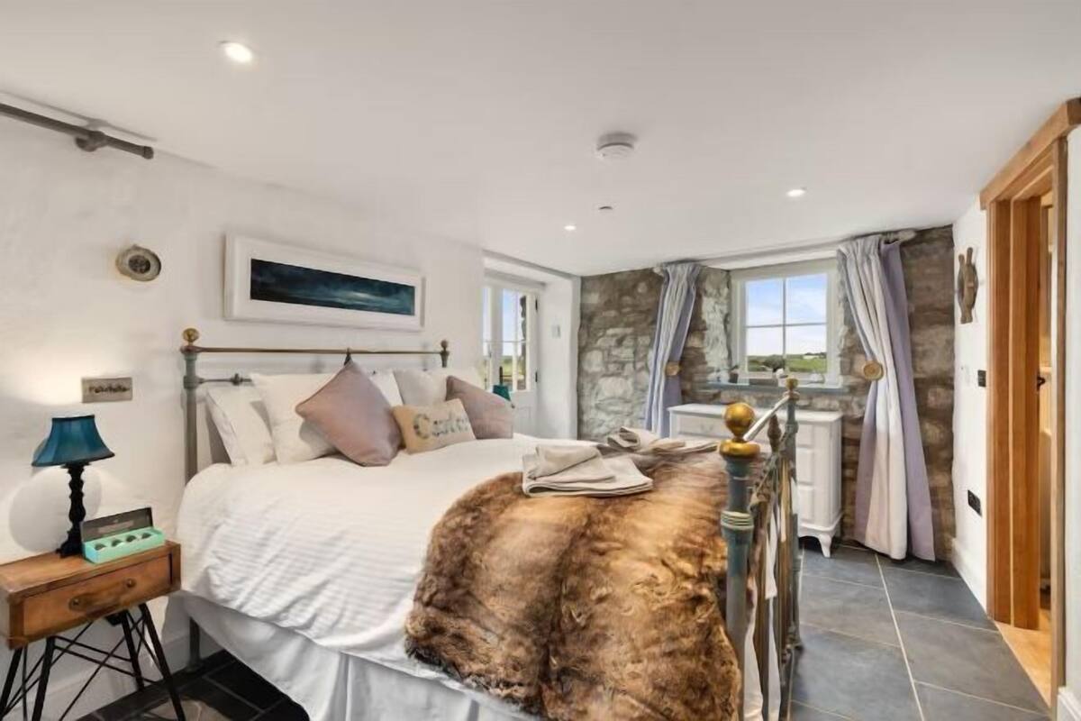 A bedroom with a bed, a nightstand, a lamp, a stone wall, a window with curtains, and a view of the outdoors.