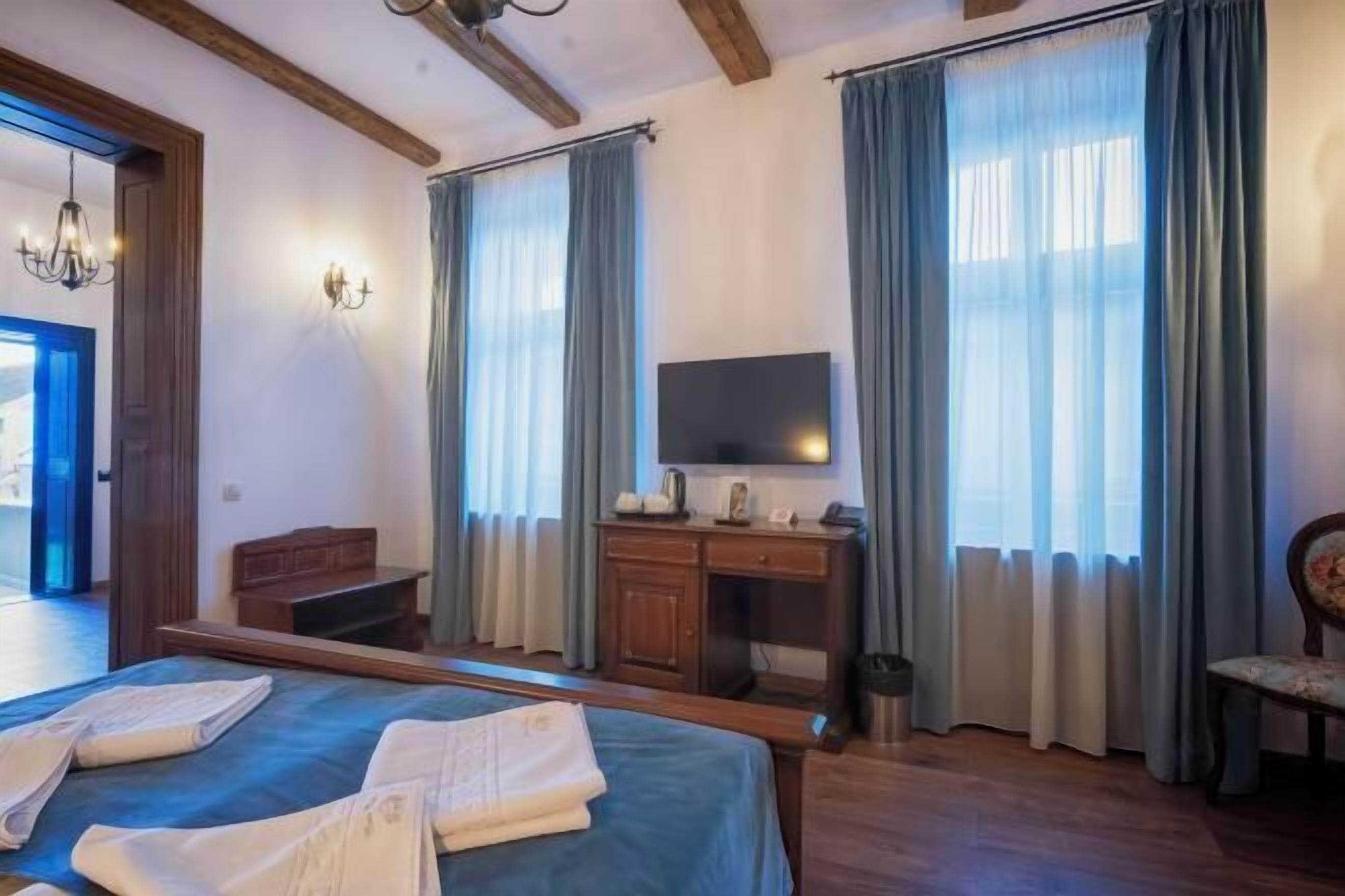 Habitación de hotel con cama, escritorio, televisor y ventanas grandes con cortinas.