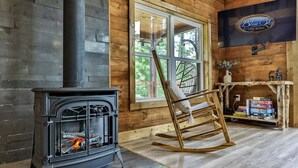 A cabin interior with a wood-burning stove, a rocking chair, a television, and a window with a view of greenery. - Logan`s Lodge | Ellijay, GA (Ellijay)