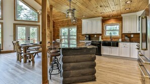 A rustic cabin interior with a wooden dining table, bar stools, a large island, and a kitchen area with white cabinets and a stainless steel refrigerator. - Logan`s Lodge | Ellijay, GA (Ellijay)