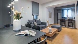 Ein modernes Wohnzimmer mit grauem Sofa, einem Couchtisch und einem Esstisch mit einer Zeitschrift. Im Bereich der Küche stehen Barhocker bereit, und ein Fenster ist mit Vorhängen versehen.