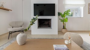 A modern living room with a fireplace, a flat-screen TV mounted above it, a comfortable armchair, and a coffee table with a magazine and candles. - Ample 3BR Apartment II in Marousi (Marousi)