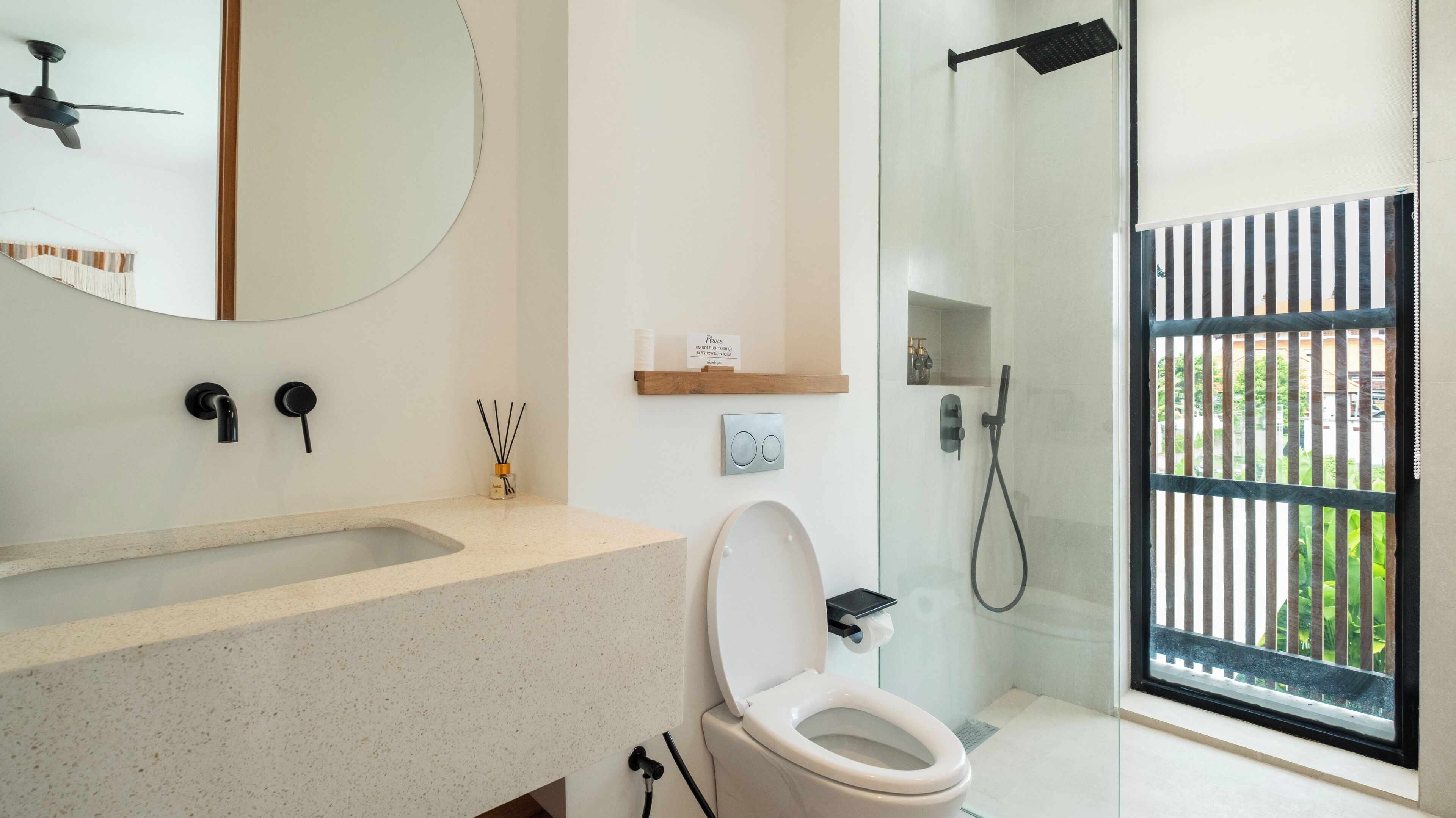 A modern bathroom with a freestanding toilet, a sink with a mirror, a shower with a glass door, and a small shelf with toiletries.