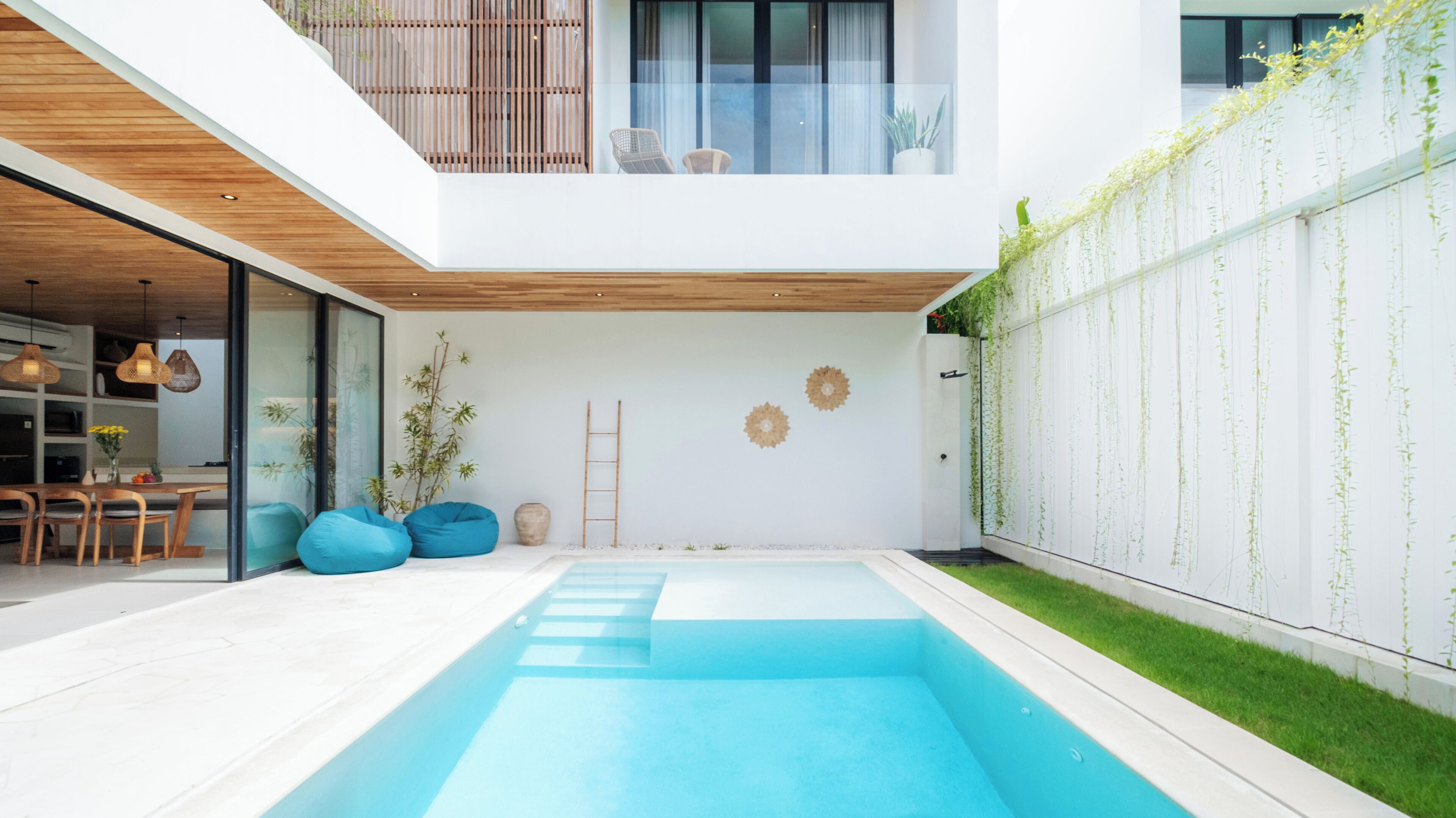 A modern house with a swimming pool, a dining area with wooden furniture, and a balcony with plants.