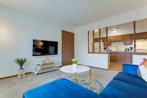 Un salon avec un téléviseur, une petite table sur laquelle est posé un vase, un canapé bleu et, au fond, une cuisine.