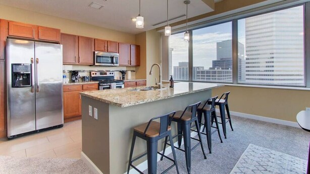 Una cocina moderna con un refrigerador de acero inoxidable, un microondas y una estufa. Hay una isla grande con taburetes y una ventana que ofrece vistas de la ciudad.